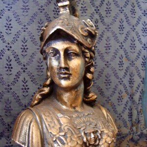May include: A gold-colored bust sculpture of a woman wearing a helmet with a decorative design. The bust is detailed with a textured, scaly chest piece and a flowing hairstyle.