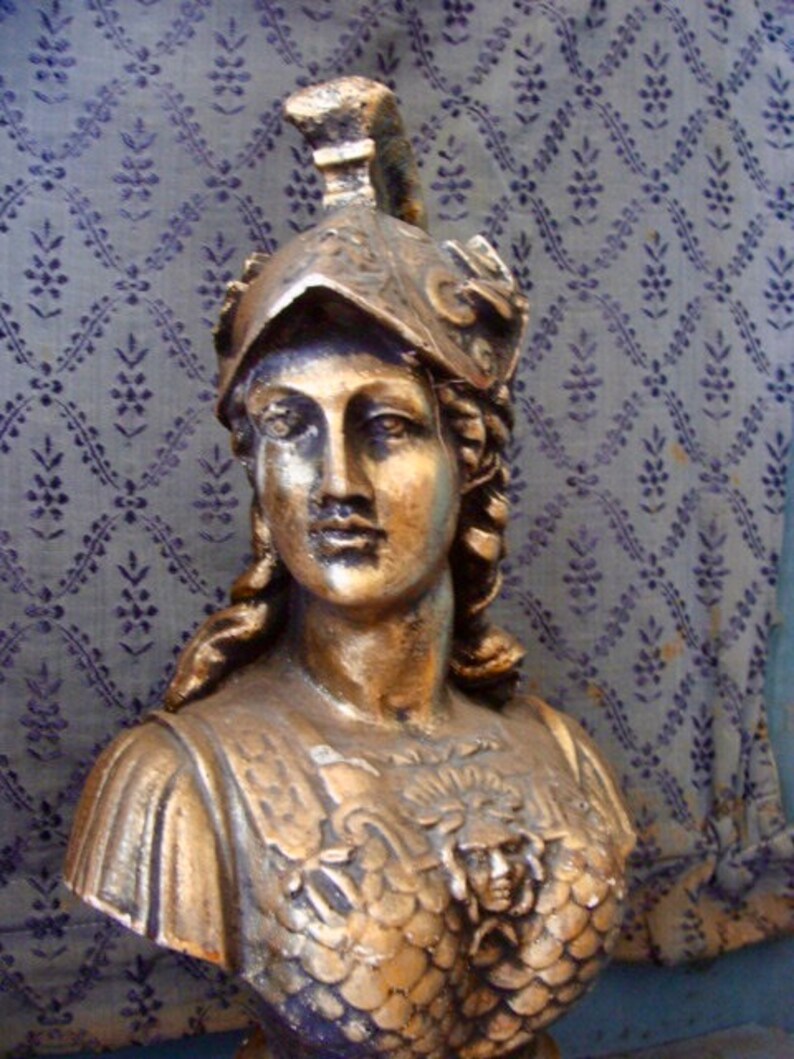 May include: A gold-colored bust sculpture of a woman wearing a helmet with a decorative design. The bust is detailed with a textured, scaly chest piece and a flowing hairstyle.