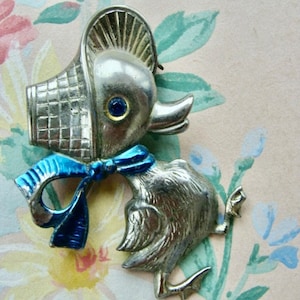 May include: A silver-toned brooch depicting a duck wearing a blue ribbon and a woven basket hat. The duck has blue eyes and a detailed, textured body.