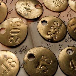May include: A collection of round, brass, numbered tags. The tags have a hole in the center and are arranged on a piece of paper with handwritten text.