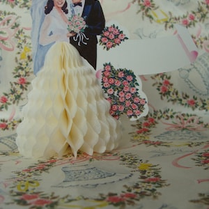 May include: A white pop-up card with a bride and groom in formal attire. The bride is wearing a white dress and veil, and the groom is wearing a black suit and tie. The card is decorated with flowers and a white ribbon.