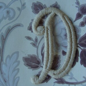 May include: A white embroidered letter D, with a decorative, cursive style, on a floral patterned background.