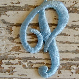May include: A light blue embroidered letter P, the letter is stitched with a satin stitch, the letter is on a white wooden background.