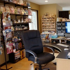 May include: A well-organized home office with a black desk, chair, and computer. Shelves are filled with jars, magazines, and craft supplies. The room has a neutral color scheme with natural light from a window.