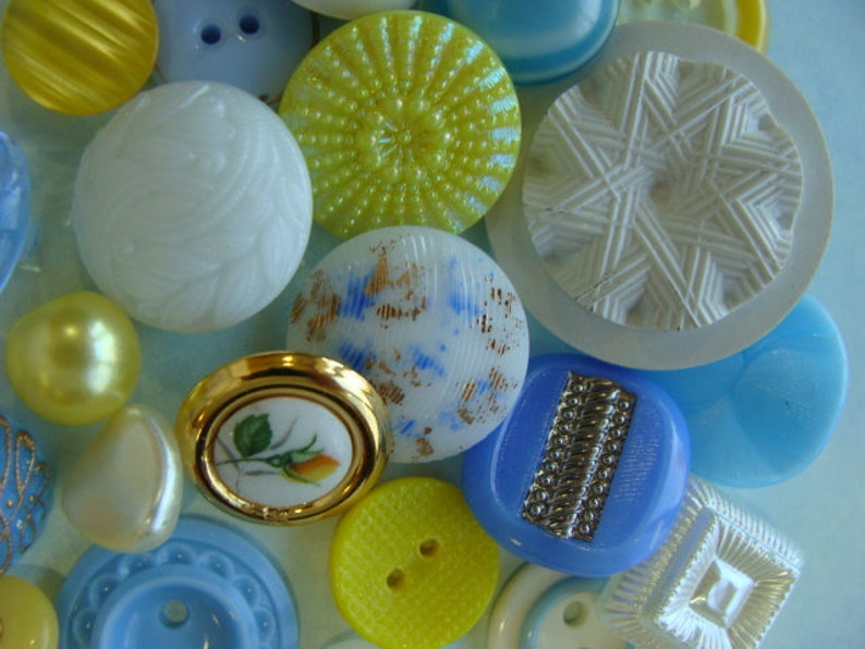 May include: Assortment of vintage buttons in various colors and patterns, including yellow, blue, white, and gold. The buttons are made of different materials, such as plastic, metal, and ceramic. Some buttons have intricate designs, while others are simple and plain.