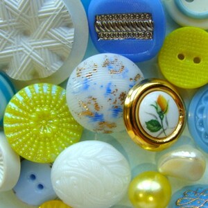 May include: Assortment of colorful buttons in various shapes and sizes, including white, blue, yellow, and gold. Some buttons have a textured surface, while others have a smooth finish. The buttons are arranged in a random pattern on a light blue background.