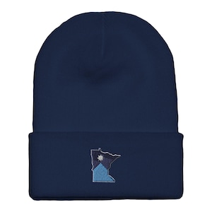 May include: A navy blue beanie hat with an embroidered design of the state of Minnesota. The design includes a star and a blue and white color scheme. The hat is made of a soft, knitted material.
