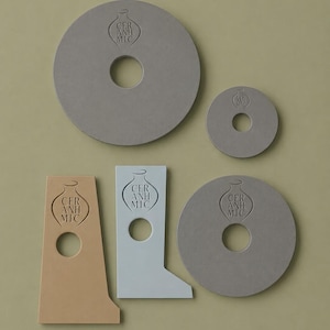May include: A collection of five flat ceramic objects in various shapes and colours. Two large grey circles, one small grey circle, one tan L-shaped piece, and one light blue L-shaped piece. Each piece features a central hole and a stamped vase logo with the text CER ANH MIC.