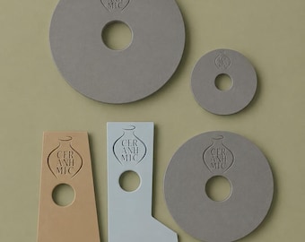 May include: A collection of five flat ceramic objects in various shapes and colors. Two large gray circles, one small gray circle, one tan L-shaped piece, and one light blue L-shaped piece. Each piece features a central hole and a stamped vase logo with the text CER ANH MIC.