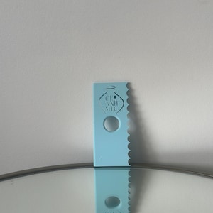 May include: A light blue plastic cake decorating tool with a scalloped edge and a hole in the centre. The tool has the text "CERAMIC" printed on it.