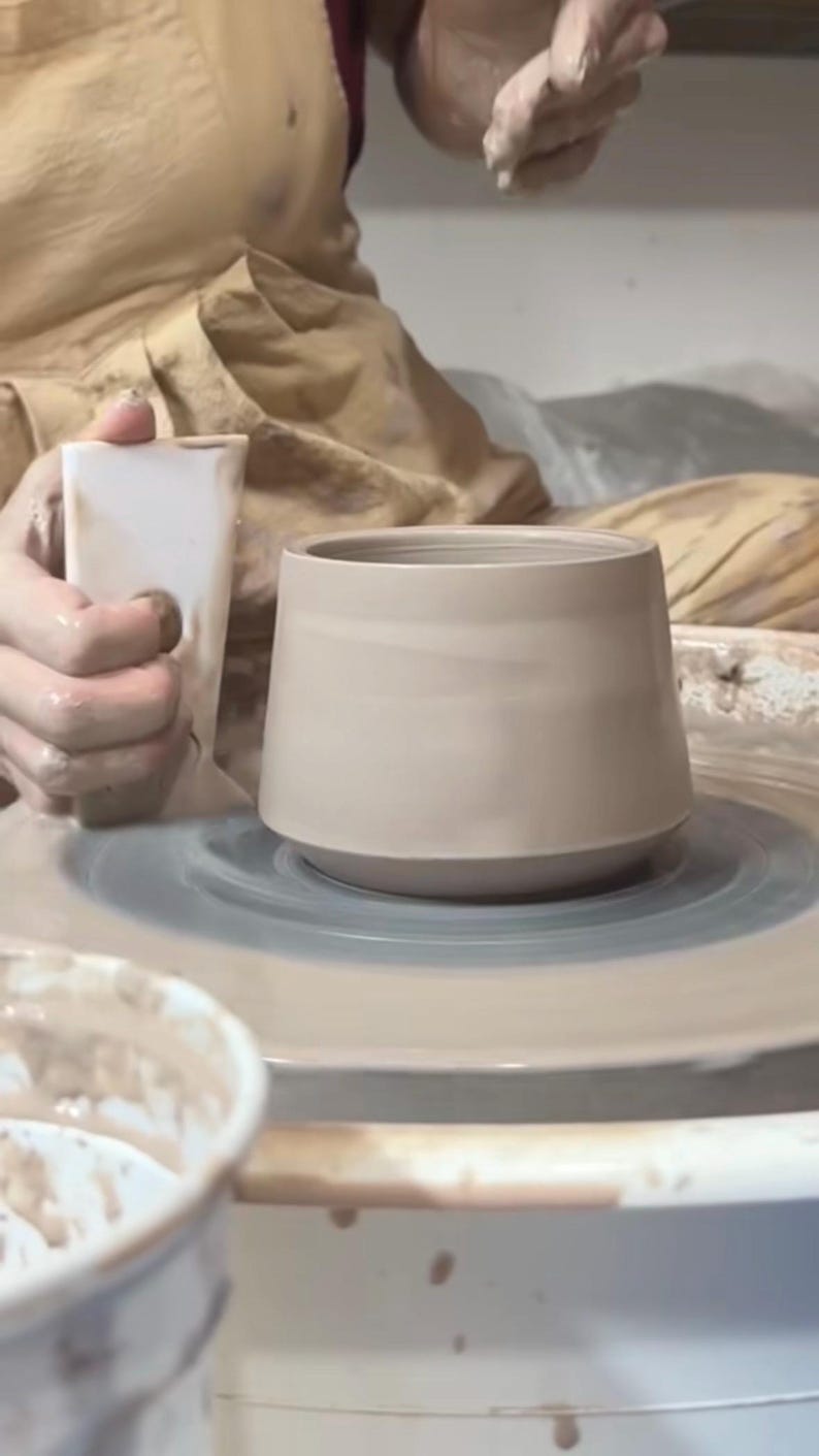 May include: A person is using a pottery wheel to shape a light brown ceramic mug. The mug is on the wheel and the person is using their hands to shape the clay.