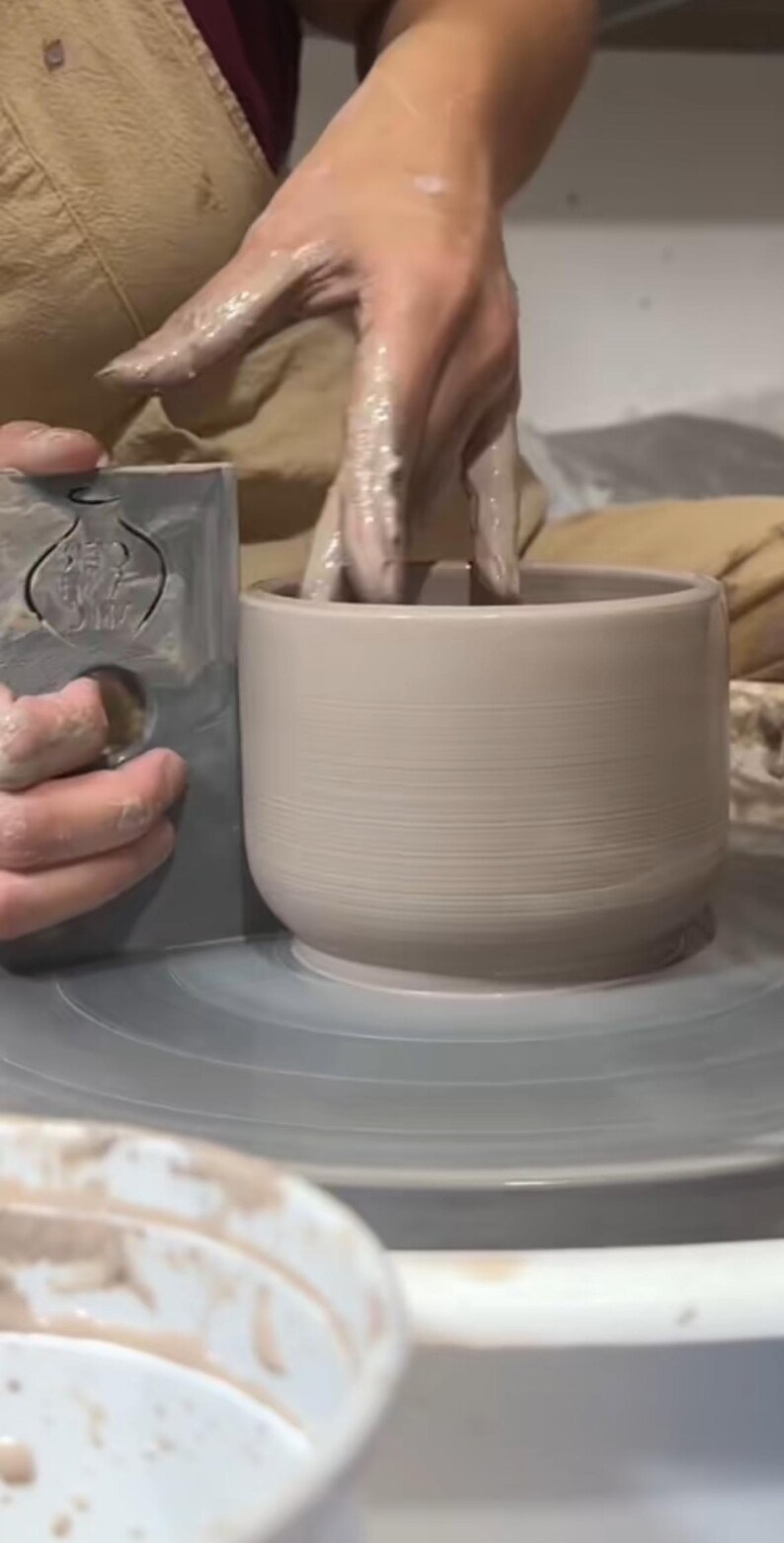 May include: A person's hands are shaping a light brown clay pot on a pottery wheel. The pot is nearly finished and has a smooth, even surface. The person is using a tool to help shape the pot.