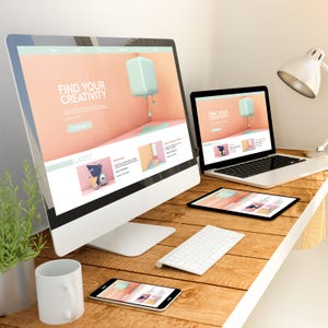 May include: A desktop workspace with a white desk and a wooden surface. There are three devices on the desk: a laptop, a tablet, and a smartphone. The laptop is open and shows a website with the text "Find Your Creativity". The tablet is also open and shows a website with the text "Find Your Creativity". The smartphone is lying flat on the desk and shows a website with the text "Find Your Creativity". There is a white keyboard and a white mug on the desk.