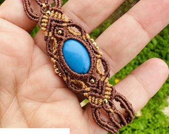 Pode incluir: Um bracelete de macramé castanho e bege com uma pedra turquesa no centro. O bracelete é amarrado com um nó e tem contas douradas.