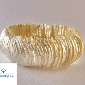 Puede incluir: Un brazalete blanco de conchas hecho de muchas conchas pequeñas que se superponen. El brazalete es un círculo y está acostado sobre una superficie blanca.