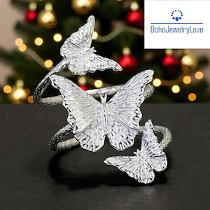 May include: Silver-coloured butterfly napkin rings. Each ring features three detailed butterfly designs. The wings are textured, and the overall design is elegant. The background is blurred with Christmas lights.