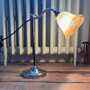 May include: A chrome and black desk lamp with a white and orange glass shade. The lamp is on and the light is shining.