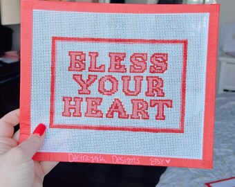 May include: A red canvas with white grid pattern featuring the words "Bless Your Heart" in red embroidered letters. The canvas is framed with a red border and has the text "Deetrajoux Designs Etsy" written in black ink at the bottom.