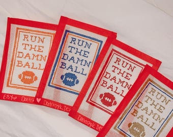 May include: Four red canvas needlepoint projects with the phrase "Run the Damn Ball" in white lettering and a football design in orange, blue, gold, and white. The projects are labeled with the Etsy shop name "Deetrazyk Designs".