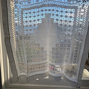 May include: A grey linen curtain with detailed lace patterns. The curtain has a central geometric design and borders with floral and geometric patterns. Sunlight filters through the delicate openwork, creating light and shadow. The curtain is hanging in a window, with a tag attached.