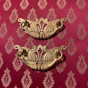 May include: Two antique brass drawer pulls with ornate, symmetrical designs. Each pull features a curved handle and decorative flourishes, ideal for vintage furniture restoration. The pulls are set against a patterned burgundy fabric.