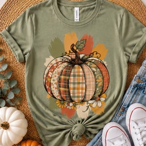 May include: An olive green short-sleeved t-shirt featuring a central graphic of a pumpkin composed of various patterned segments, including plaid and floral prints. The pumpkin is accented with brushstroke-style background colours and small flowers.