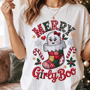 May include: A white t-shirt featuring a graphic of a ghost inside a patchwork Christmas stocking. The text reads MERRY GIRLY BOO. The design includes red and green candy canes, snowflakes, holly leaves, and small gold stars in a festive style.
