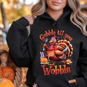 May include: Black hoodie with a Thanksgiving-themed graphic. The design features a cartoon turkey holding a fork and knife, with the words "Gobble til you Wobble". The artwork is set against a fall-themed background. The hoodie is made of a soft material.