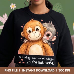 Puede incluir: Sudadera negra con una ilustración de dibujos animados de dos monos abrazándose. Los monos son marrones y grises con ojos grandes y caras sonrientes. El texto "It's okay not to be okay... you're not alone" está impreso debajo.