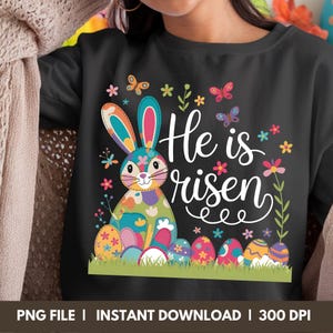 May include: A black t-shirt featuring a colourful Easter bunny and the text "He is risen" in white. The bunny is surrounded by flowers and Easter eggs.