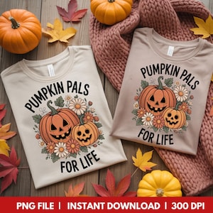 Puede incluir: Dos camisetas beige con el texto "PUMPKIN PALS FOR LIFE" y un diseño de dos calabazas sonrientes rodeadas de flores. La imagen incluye calabazas, hojas de otoño y una bufanda rosa sobre una superficie de madera.