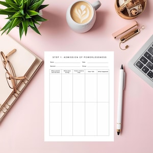 Op de afbeelding: Een wit werkblad met de tekst "STEP 1: ADMISSION OF POWERLESSNESS" ligt op een roze oppervlak. Het werkblad heeft secties om over alcoholgebruik te schrijven. Een witte pen, een laptop en een kopje koffie liggen ook op het oppervlak.