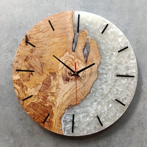 May include: A round wooden and white resin clock with black hands and numbers. The wood has a natural grain pattern and the resin has a marbled effect.