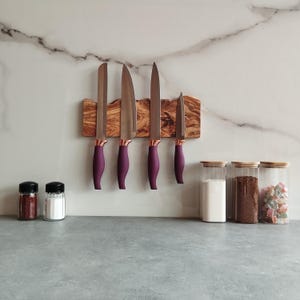 Puede incluir: Cinco cuchillos de cocina plateados con mangos morados están montados en una tira magnética de madera. La tira está unida a una pared de azulejos blancos.