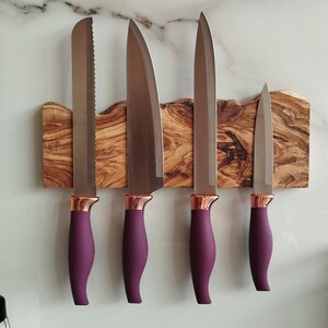 Pu&ograve; includere: Quattro coltelli da cucina con manici viola e dettagli in oro rosa sono montati su un portacoltelli magnetico in legno. Il supporto &egrave; in legno di ulivo con venature del legno naturali.
