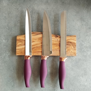 Pu&ograve; includere: Tre coltelli da cucina con manici viola e dettagli in oro rosa sono montati su un portacoltelli magnetico in legno.