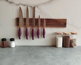 Può includere: Un portacoltelli magnetico in legno con sei coltelli. I coltelli hanno lame argentate, rinforzi color oro rosa e manici color prugna. Sono visibili anche due piccoli barattoli di spezie e tre barattoli di vetro con coperchi in legno.