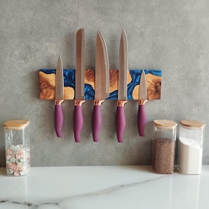 May include: A set of six kitchen knives with purple handles and copper-colored bolsters, mounted on a wooden magnetic knife holder with blue resin accents. Jars with wooden lids are on the counter.