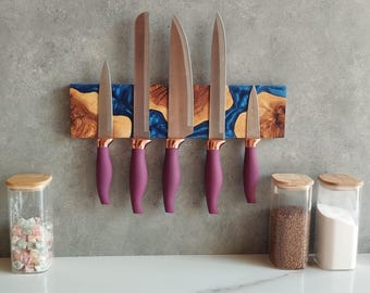Può includere: Un set di sei coltelli da cucina con manici viola e dettagli color rame, montati su un portacoltelli magnetico in legno con accenti di resina blu. Barattoli con coperchi in legno sul bancone.