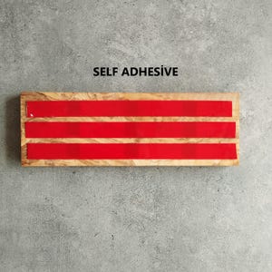 Pu&ograve; includere: Un blocco di legno con tre strisce autoadesive rosse. Il blocco ha una venatura del legno naturale. Il testo "SELF ADHESIVE" &egrave; stampato sopra il blocco.