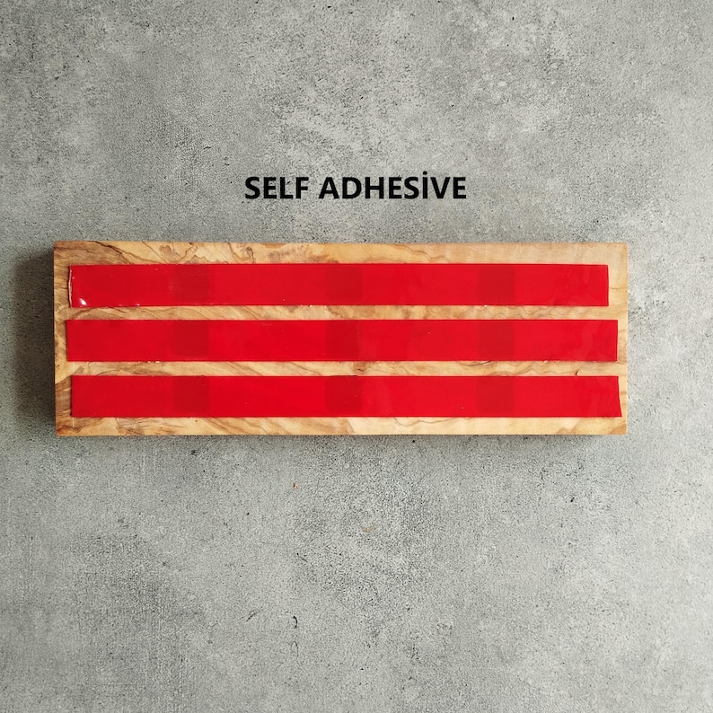 Pu&ograve; includere: Un blocco di legno con tre strisce autoadesive rosse. Il blocco ha una venatura del legno naturale. Il testo "SELF ADHESIVE" &egrave; stampato sopra il blocco.