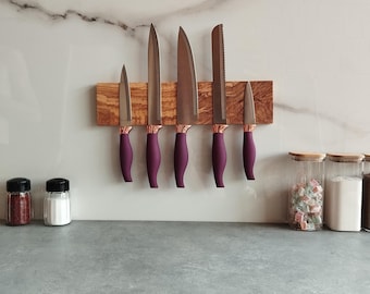 Può includere: Un portacoltelli magnetico in legno con sei coltelli con manici viola e dettagli in oro rosa. I coltelli sono fissati al supporto su una parete piastrellata bianca.