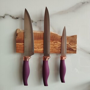 Pu&ograve; includere: Tre coltelli da cucina con manici viola e dettagli in oro rosa sono montati su una striscia magnetica in legno. La striscia &egrave; realizzata in legno di ulivo e ha una finitura naturale e rustica.