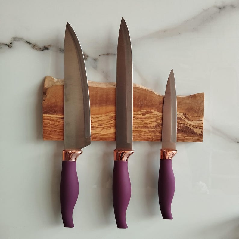 Pu&ograve; includere: Tre coltelli da cucina con manici viola e dettagli in oro rosa sono montati su una striscia magnetica in legno. La striscia &egrave; realizzata in legno di ulivo e ha una finitura naturale e rustica.