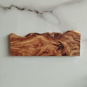 Pu&ograve; includere: Un tagliere in legno naturale con un bordo ondulato e una texture marrone e granulosa. Il tagliere &egrave; in legno di ulivo.