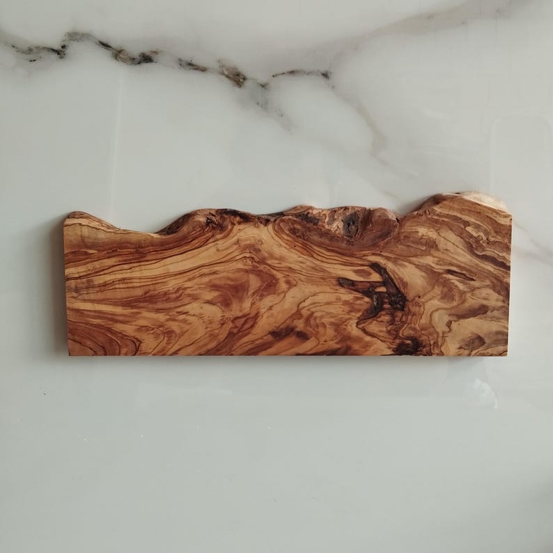 Pu&ograve; includere: Un tagliere in legno naturale con un bordo ondulato e una texture marrone e granulosa. Il tagliere &egrave; in legno di ulivo.