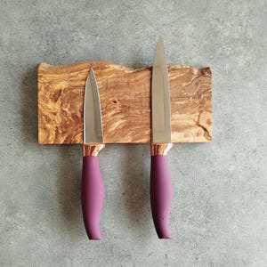 Pu&ograve; includere: Due coltelli da cucina con manici viola sono montati su un portacoltelli magnetico in legno. Il supporto &egrave; in legno di ulivo e ha una finitura naturale e rustica.