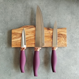 Pu&ograve; includere: Tre coltelli da cucina con manici viola sono montati su una striscia magnetica in legno. I coltelli sono disposti in ordine di dimensione, con il coltello pi&ugrave; grande a sinistra e il coltello pi&ugrave; piccolo a destra.