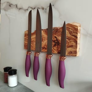 Pu&ograve; includere: Un set di quattro coltelli da cucina con manici viola e dettagli in oro rosa sono montati su un portacoltelli magnetico in legno. Il supporto &egrave; in legno di ulivo e presenta una venatura del legno naturale.