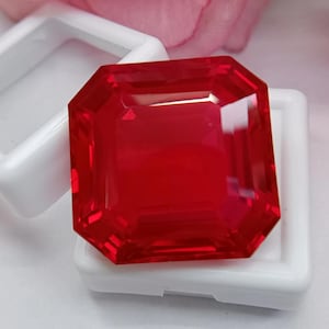 May include: A vibrant, square-cut red gemstone with faceted edges, displaying a deep crimson hue and high clarity. The stone is positioned resting on a white plastic display case, which features a square frame and a clear, flexible membrane insert.